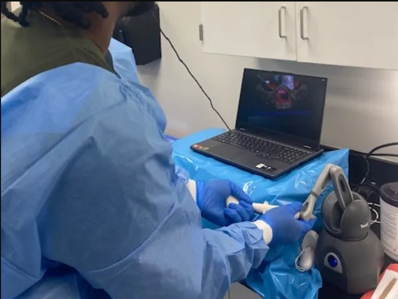 A surgeon in scrubs uses the SurgicalEd VR haptic console to practice a procedure against an anatomical model on screen.